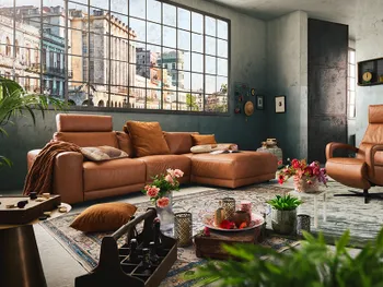 Großzügiges Wohnzimmer mit cognacfarbenem Ledersofa und passendem Sessel im Industrial-Look, dekoriert mit bunten Kissen, Blumen und orientalischen Accessoires vor einer großen Fensterfront mit Stadtblick.